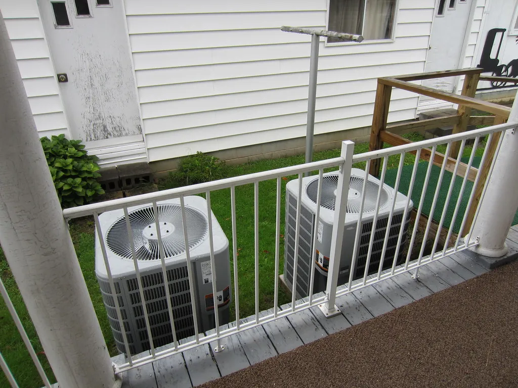 Two central air condensers behind a porch railing beside a house with white siding